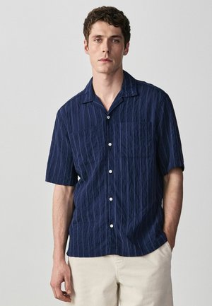 Young man standing with hand in pocket, wearing a navy blue short-sleeve button-up shirt and light beige pants against a plain background.