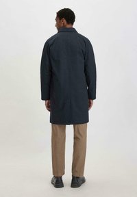 Marineblauer Mantel mit strukturiertem Design, hoher Kragen und geradem Schnitt. Kombiniert mit beigen Hosen und schwarzen Schuhen. Glatte Textur.