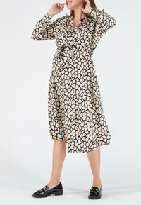 Cremefarbenes Kleid mit einem schwarzen abstrakten Muster, Knopfleiste, langen Ärmeln, einem Gürtel in der Taille und knielangem Saum. Getragen mit schwarzen Slippers.