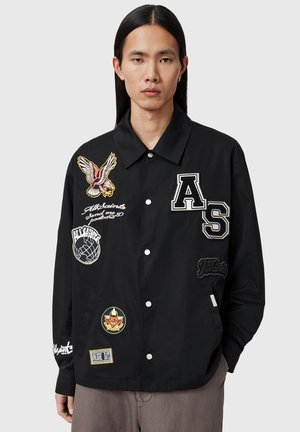 Ung mand med langt sort hår iført en sort knappet jakke dekoreret med flere mærker og logoer, stående foran en ensfarvet baggrund.