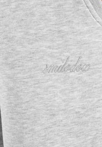 Hellgrauer Stoff mit subtiler Stickerei des Wortes "smilodosa" in passendem Garn near der oberen Kante.