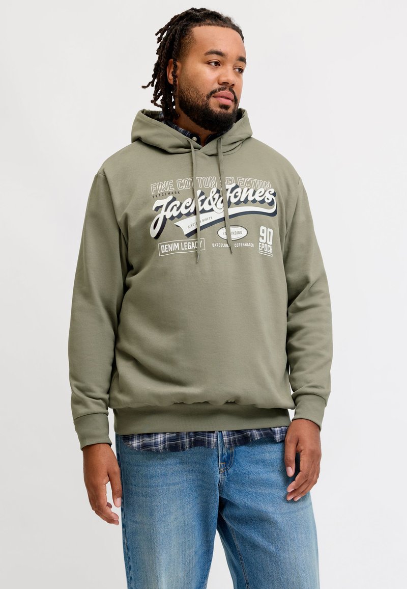 Homme avec barbe et dreadlocks portant un sweat à capuche Jack & Jones vert olive et un jean bleu, regardant sur le côté sur un fond clair uni.