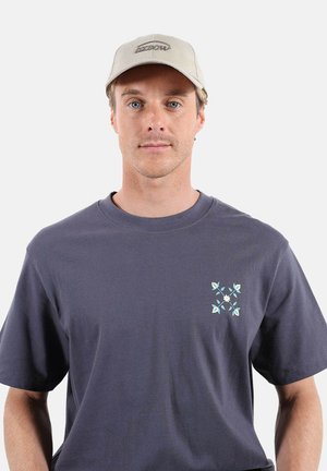 Hombre con gorra beige y camiseta gris oscuro con un pequeño diseño floral azul y blanco en el lado izquierdo del pecho, fondo neutro.