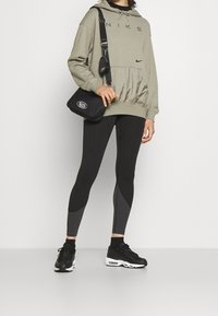 Olivově zelená mikina Nike s černými akcenty, spárovaná s černými a šedými legínami. Černé tenisky a malá černá crossbody taška dotvářejí celkový vzhled.