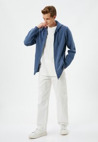 Sudadera azul con cremallera y cordones, llevada sobre una camiseta blanca, combinada con pantalones cargo blancos y zapatillas blancas. Fondo liso.