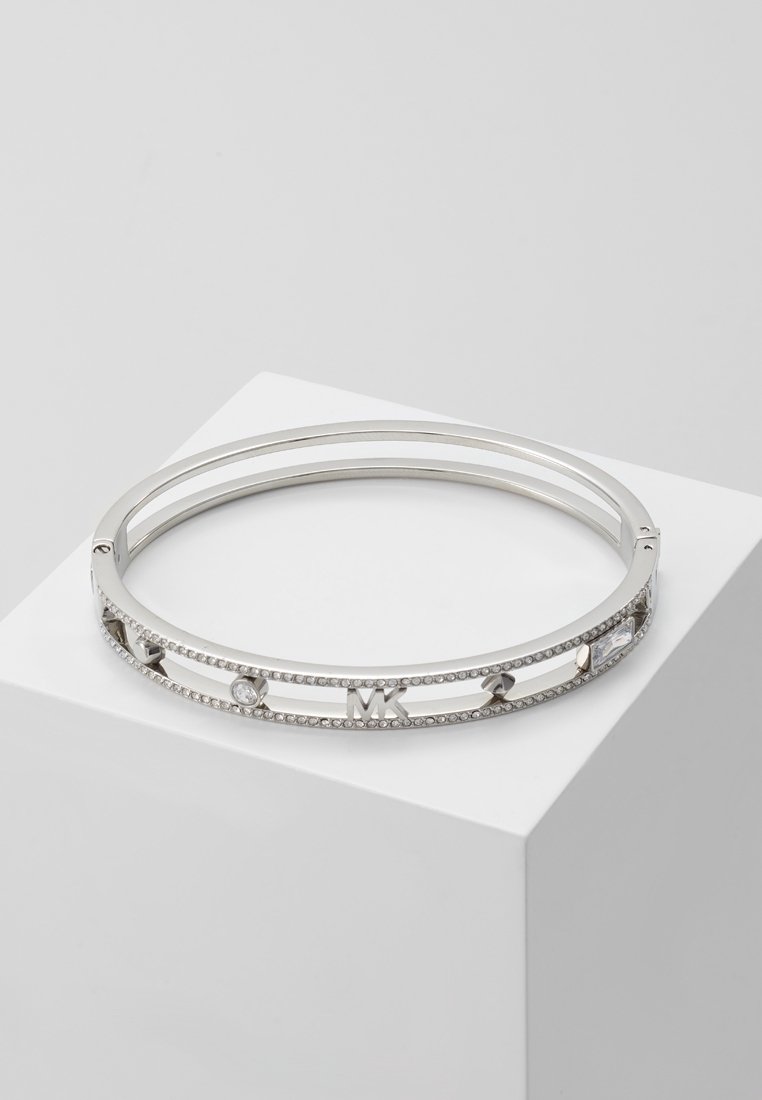 Bracelet jonc ton argent avec cristaux clairs et accents logo MK, présenté sur un socle blanc incliné sur fond gris.