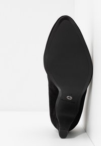 Chaussure à talon haut en daim noir avec bout pointu, texture lisse et semelle en caoutchouc plate. Présente un logo métallique près du talon. Taille 37.