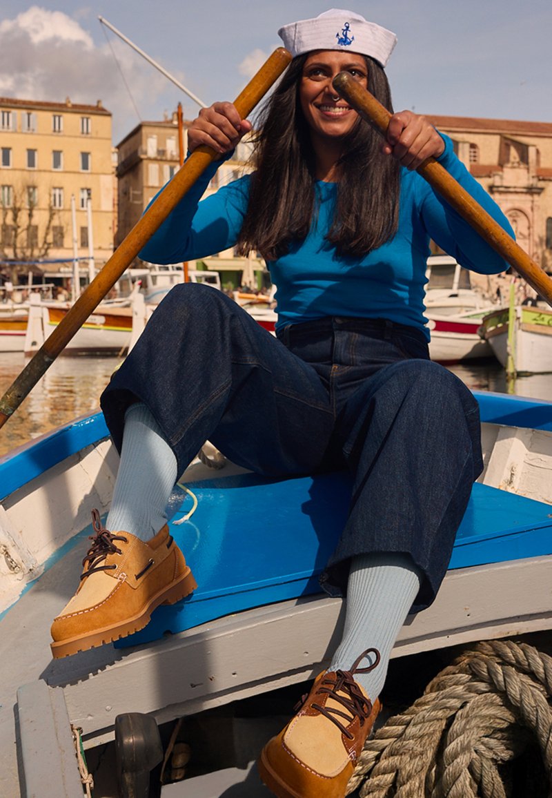 Jeune femme portant un chapeau de marin rame un petit bateau dans un port ensoleillé avec des bâtiments et des bateaux en arrière-plan.