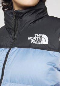 Kamizelka z pikowanymi panelami, z czarną górną częścią i jasnoniebieską dolną częścią. Posiada wysoki kołnierz oraz zamek błyskawiczny z przodu. Logo jest wyraźnie widoczne.