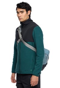 Giacca in pile con parte superiore nera e parte inferiore teal, apertura con zip, tasche laterali e una tracolla grigia regolabile.