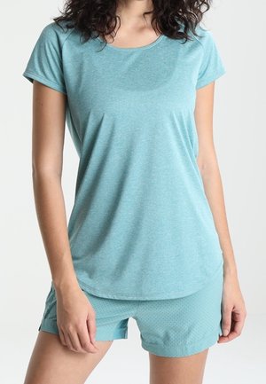 Frau trägt ein hellblaues T-Shirt mit kurzen Ärmeln und passende strukturierte Shorts, stehend vor einem schlichten weißen Hintergrund.