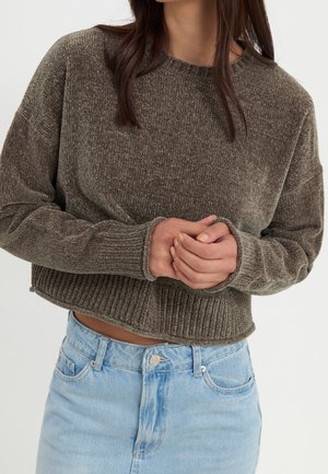 Femme portant un pull court texturé vert olive avec poignets et ourlet côtelés, associé à un jean taille haute bleu clair, mains jointes.