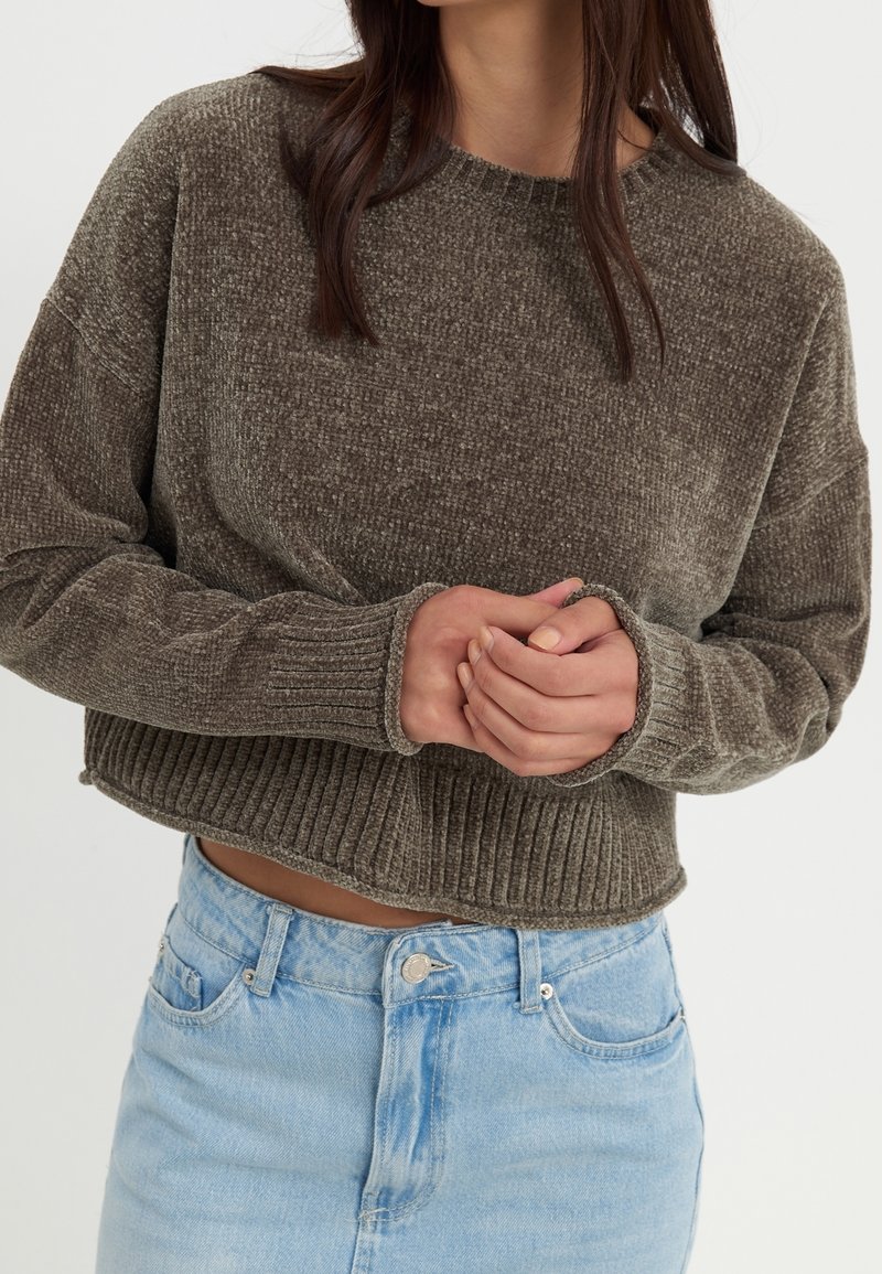 Femme portant un pull court texturé vert olive avec poignets et ourlet côtelés, associé à un jean taille haute bleu clair, mains jointes.