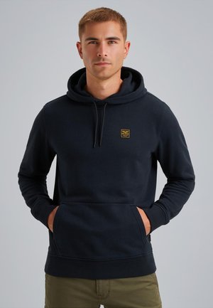 Giovane uomo che indossa un hoodie nero con tasca frontale e pantaloni khaki, in piedi con le mani nelle tasche su uno sfondo neutro.