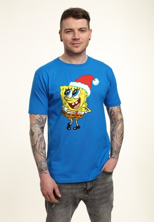 Niebieska bawełniana koszulka z kreskówkową postacią w czapce Mikołaja, intensywnie żółty kolor i czarne buty. Widoczna wytatuowana ręka.