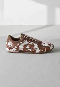 Kengät, joissa on lehmäntilkut ruskeana ja valkoisena, mokkanahkakoristuksilla, pyöreällä varvalla ja beigeillä nauhoilla. Litteä kumipohja, jossa on teksturoidut yksityiskohdat.