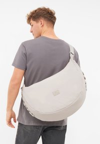 Borsa a tracolla grigia chiara realizzata in tessuto morbido con forma arrotondata, tracolla regolabile e una discreta toppa con logo sul davanti.