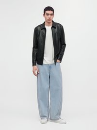 Young man wearing a black leather jacket, white t-shirt, light blue wide-leg jeans, and white sneakers standing against a plain background.
