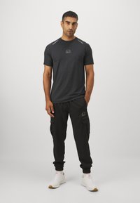 Männer T-Shirt in Grau mit kurzen Ärmeln, runder Ausschnitt und reflektierenden Akzenten; kombiniert mit schwarzen Cargo-Hosen mit Taschen und elastischen Bündchen.