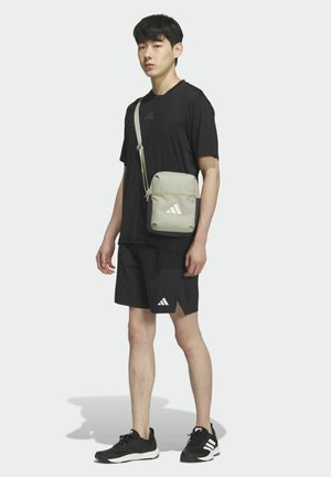 Šedá kabelka přes rameno se zipovým uzávěrem, zdobená bílými logo akcenty. Model má na sobě černé tričko, šortky a černé sportovní boty.