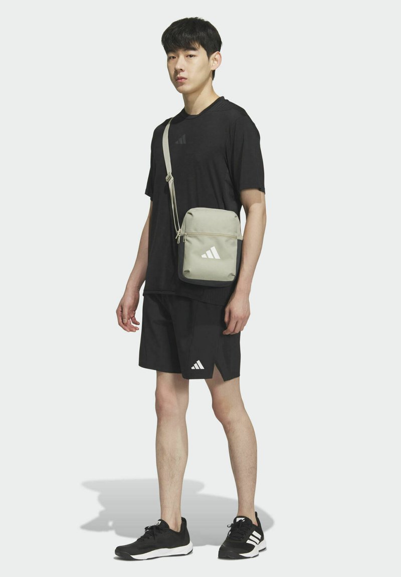 Sivá crossbody taška so zipsovým uzáverom, so bielymi logovými akcentmi. Model nosí čierne tričko, šortky a čierne športové topánky.