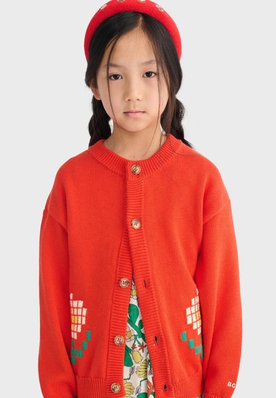 Niña joven con el cabello trenzado que lleva un cárdigan rojo con patrones florales y una boina roja a juego sobre un fondo liso.