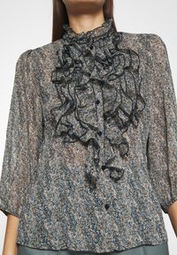 Blusa transparente y estampada con cuello y puños con volantes. Presenta una mezcla de colores azul, beige y negro, y cierre de botones en la parte frontal.
