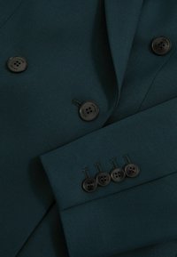 Tumši zilais žakets ar teksturētu audumu, dubultās krūškurves dizainu, melniem pogām un četrām akcenta pogām uz piedurknes.