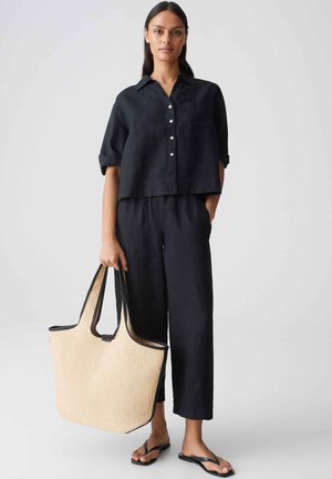 Femme portant une chemise noire à boutons et un pantalon, tenant un sac fourre-tout tissé beige, debout pieds nus sur fond gris.