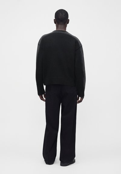 Personne debout, vue de dos, portant un pull noir avec des rayures grises sur les manches et un pantalon noir ample devant un fond uni.