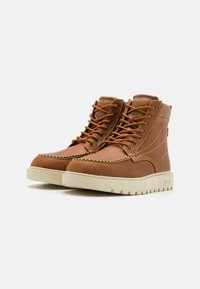 Botas de cuero marrón hasta el tobillo con suelas de goma crema, que cuentan con un diseño de cordones, detalles de costura y un cuello acolchado para mayor soporte.