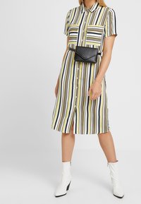 Pruhované košilové šaty v žluté, navy a bílé barvě s krátkým rukávem, dvěma kapsami na prsou a předním rozparkem. Doplněné černou ledvinkou.