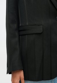 Mustra mustritäck blazer, mille vasakul küljel on varrukate ja klapi tasku detail, kantud helesiniste denim teksade üle, käsi pingevabalt külje juures.