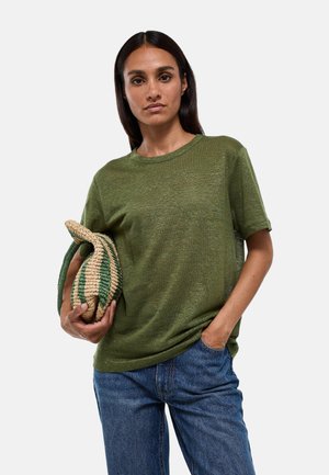 Femme aux cheveux bruns raides portant un haut vert à manches courtes et un jean bleu tient un sac tissé vert et beige sur un fond uni.