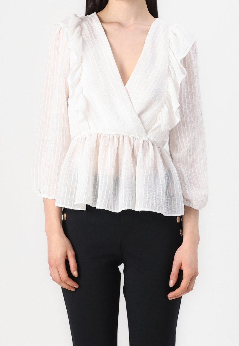 Femme portant un chemisier blanc texturé avec des volants, un col en V profond et des manches longues transparentes, associé à un pantalon noir.