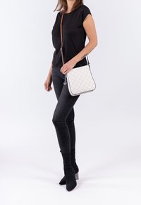 Bílá prošívaná kabelka přes rameno se stříbrnými detaily, která má hnědý popruh a zaoblené rohy, je nošena modelem v černém oblečení a džínách.