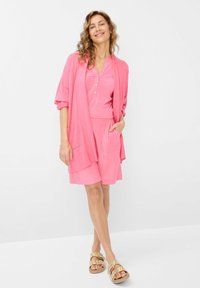 Roze, lichtgewicht, kortemouwtop met knopen, gecombineerd met een bijpassende rok tot de knie en een lange cardigan; sandalen met gevlochten accenten.