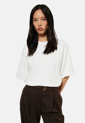 Kobieta z długimi ciemnymi włosami, ubrana w białą koszulę z krótkim rękawem z małymi ozdobami oraz ciemne spodnie z wysokim stanem i paskiem.