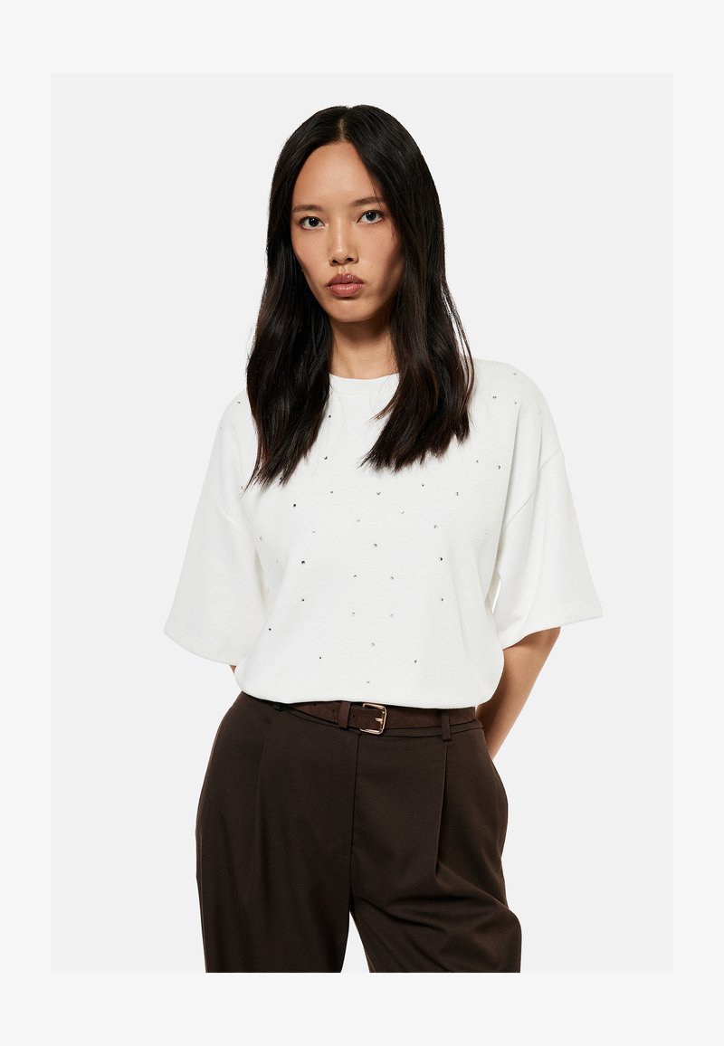 Mujer con cabello largo y oscuro, vistiendo una camisa blanca de manga corta con pequeños adornos y pantalones oscuros de talle alto con cinturón.