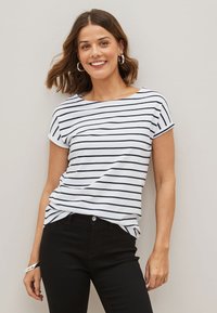 Femme souriante aux cheveux bruns mi-longs porte des boucles d'oreilles créoles argentées, une chemise blanche à manches courtes avec des rayures noires et un pantalon noir.