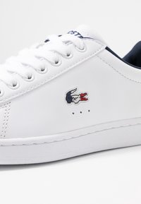 Baskets en cuir blanc avec bout rond, lacets plats blancs, intérieur bleu marine, logo brodé en bleu, rouge et blanc, et détails de couture visibles.