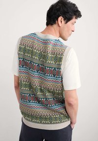 Mann, der eine mehrfarbige, gemusterte Strickweste über einem weißen kurzärmeligen Hemd trägt, mit dem Rücken zur Kamera und den Händen in den Taschen vor einem einfarbigen Hintergrund.