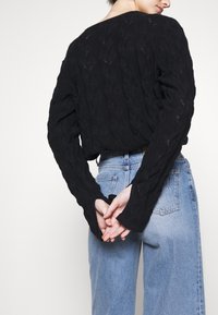 Persona vestita con un maglione nero a trecce e jeans azzurri, in piedi con le mani incrociate dietro la schiena.