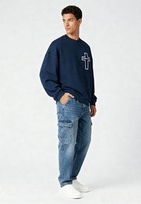 Jeune homme portant un sweat-shirt bleu marine avec un motif croisé "Karma Kills", un jean cargo bleu et des baskets blanches, debout avec une main dans la poche.