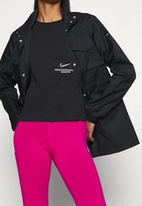 Svart zip-up jacka med tryckknappar och platta fickor, tillsammans med en svart t-shirt med en vit logotyp, samt klara rosa leggings.