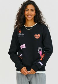 Čierna mikina so širokou škálou farebných nášiviek, vrátane červeného srdca, kvetov a textu. Má okrúhly výstrih a rebrované manžety.