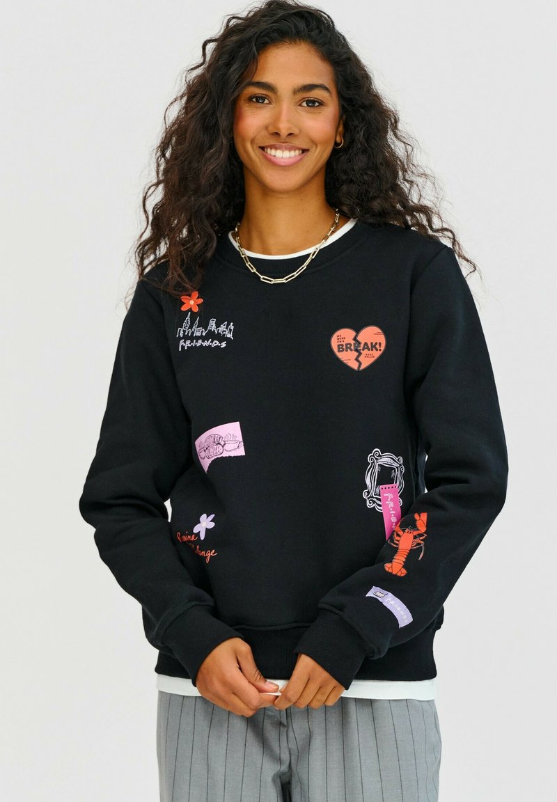 Čierna mikina so širokou škálou farebných nášiviek, vrátane červeného srdca, kvetov a textu. Má okrúhly výstrih a rebrované manžety.