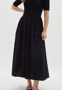 Schwarzes Kleid mit einem taillierten, gerippten Oberteil und einem fließenden, gerafften Rock. Sanfte Textur, knöchellang, mit kurzen Ärmeln und minimaler Detailierung.