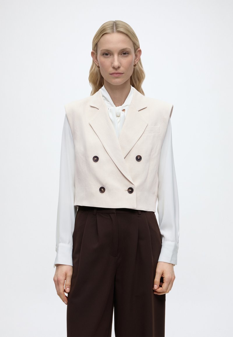 Γυναίκα με ξανθά μαλλιά που φορά ένα λευκό διπλόστενο γιλέκο χωρίς μανίκια πάνω από μια λευκή μπλούζα με μακριά μανίκια και σκούρο καφέ παντελόνια ψηλόμεσα.