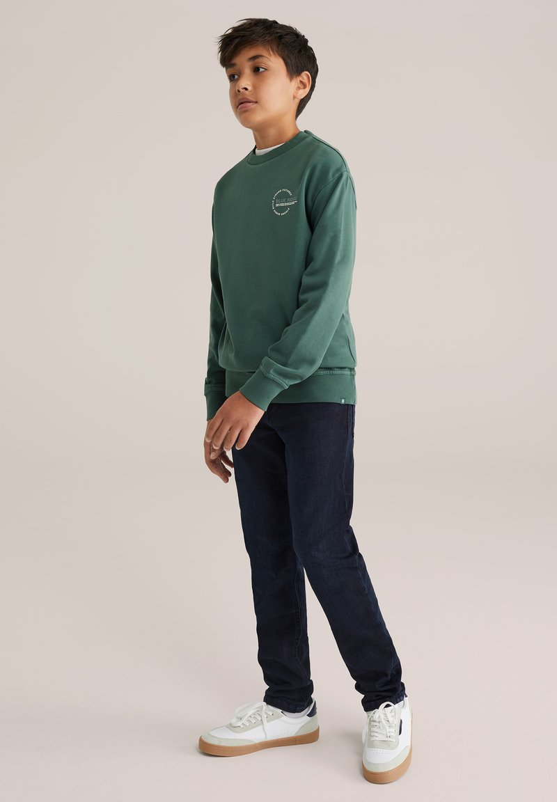 Zelená bavlněná mikina s kulatým výstřihem a Royal lano rukávy, kombinovaná s tmavě modrými džíny a bílými teniskami se světle hnědými podrážkami.